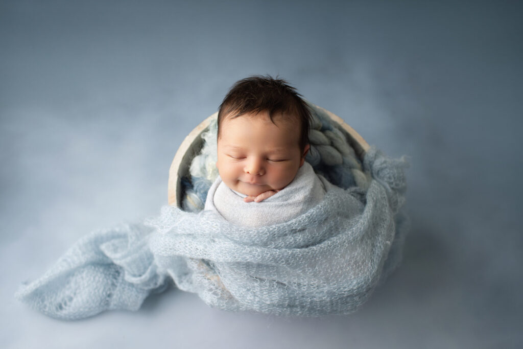 Newborn baby wrapped in a light blue blanket, sleeping inside a round basket on a soft blue background.