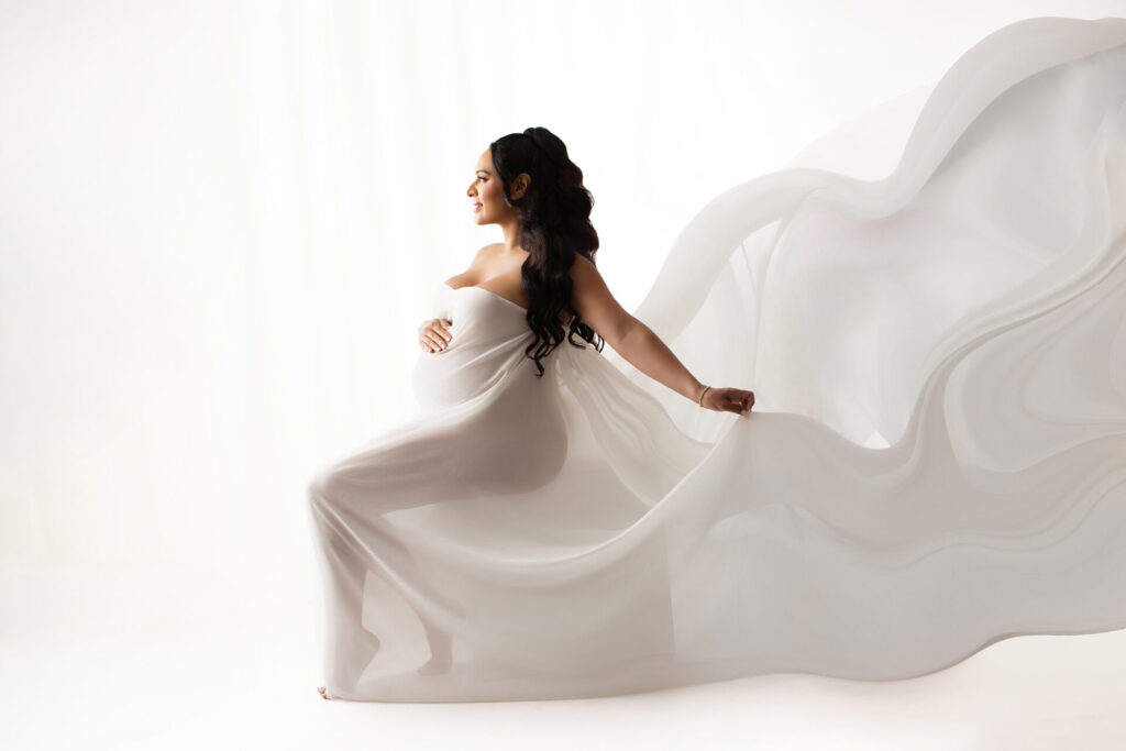 Pregnant woman in a sheer white gown sits sideways, holding her belly, with flowing fabric extending behind her against a white background.
