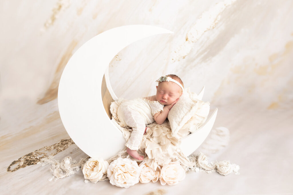 Newborn baby sleeping on a crescent moon prop, surrounded by white flowers and soft fabrics, against a neutral background.