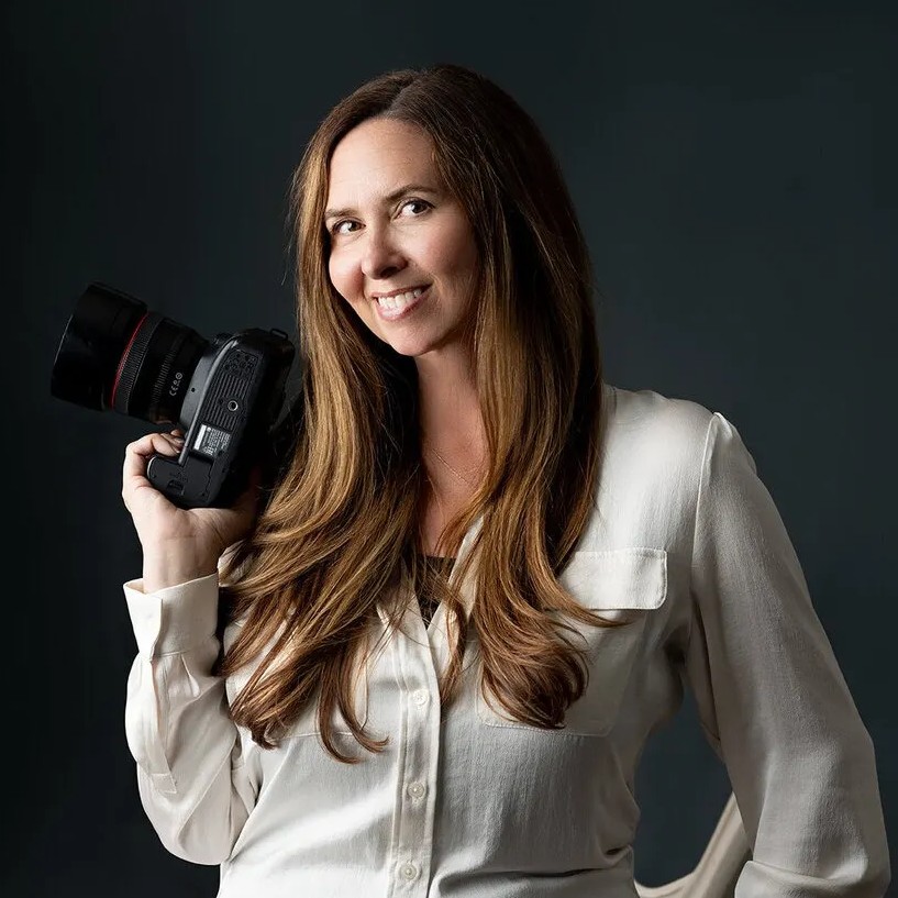 Woman with long brown hair wearing a white shirt smiles and holds a professional camera on her shoulder against a dark background.