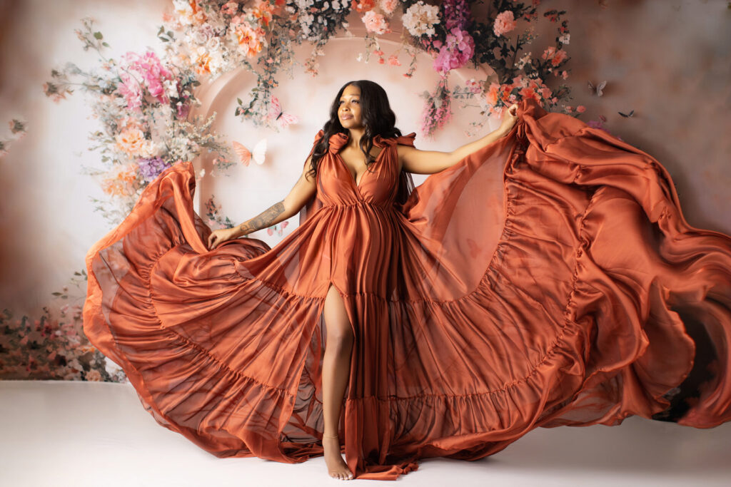 Woman in a flowing rust-colored dress poses with arms outstretched in front of a floral backdrop.