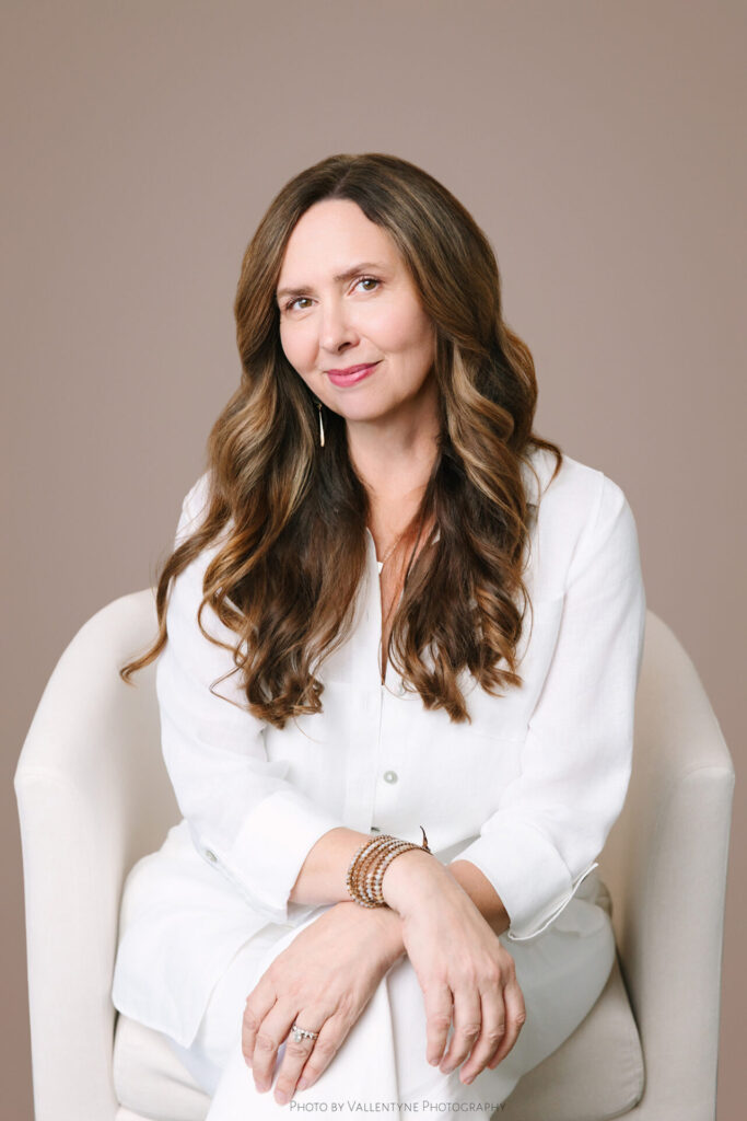 A woman with long brown hair, wearing a white outfit, sits on a light-colored chair against a plain background, hands resting on her knee.