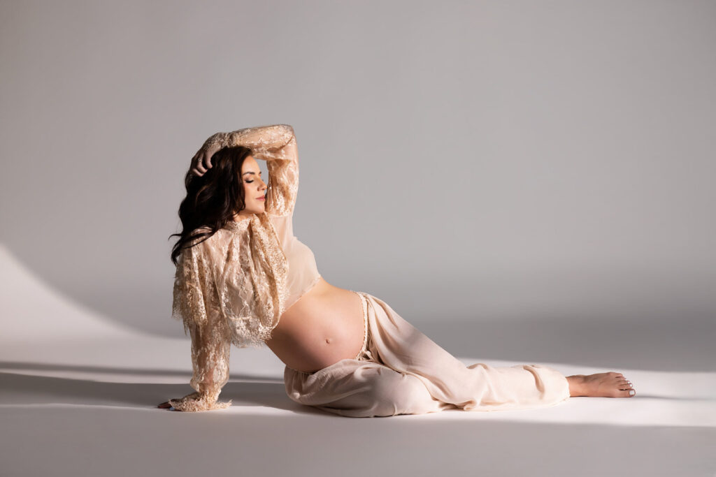 Pregnant woman in a beige lace top and flowing pants reclining on the floor.